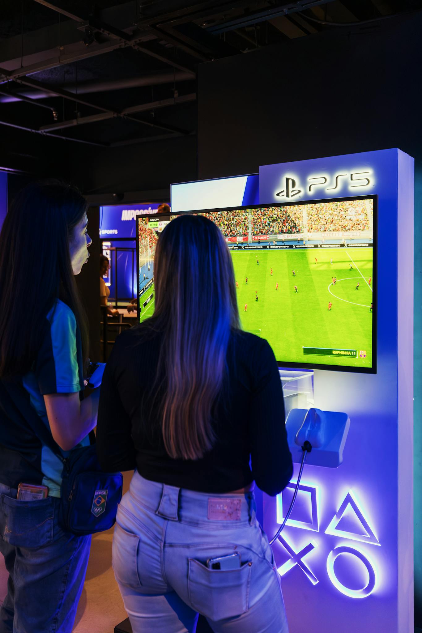 Two women playing FIFA on a PlayStation 5 console in a modern gaming store.